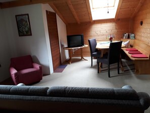 A cozy room with a wooden ceiling, a television, a dining table with chairs, a sofa, and a bench with red cushions.