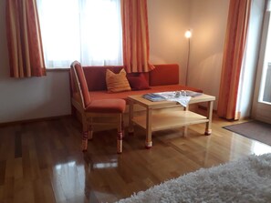 Ein gemütliches Zimmer mit Holzboden, einem Sofa mit orangefarbenen Kissen, einem kleinen Tisch und einer Lampe.