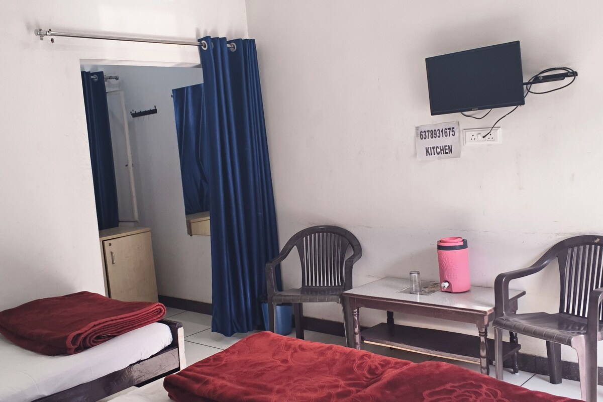 A hotel room with two beds, a desk with chairs, a television, and a kitchenette area.