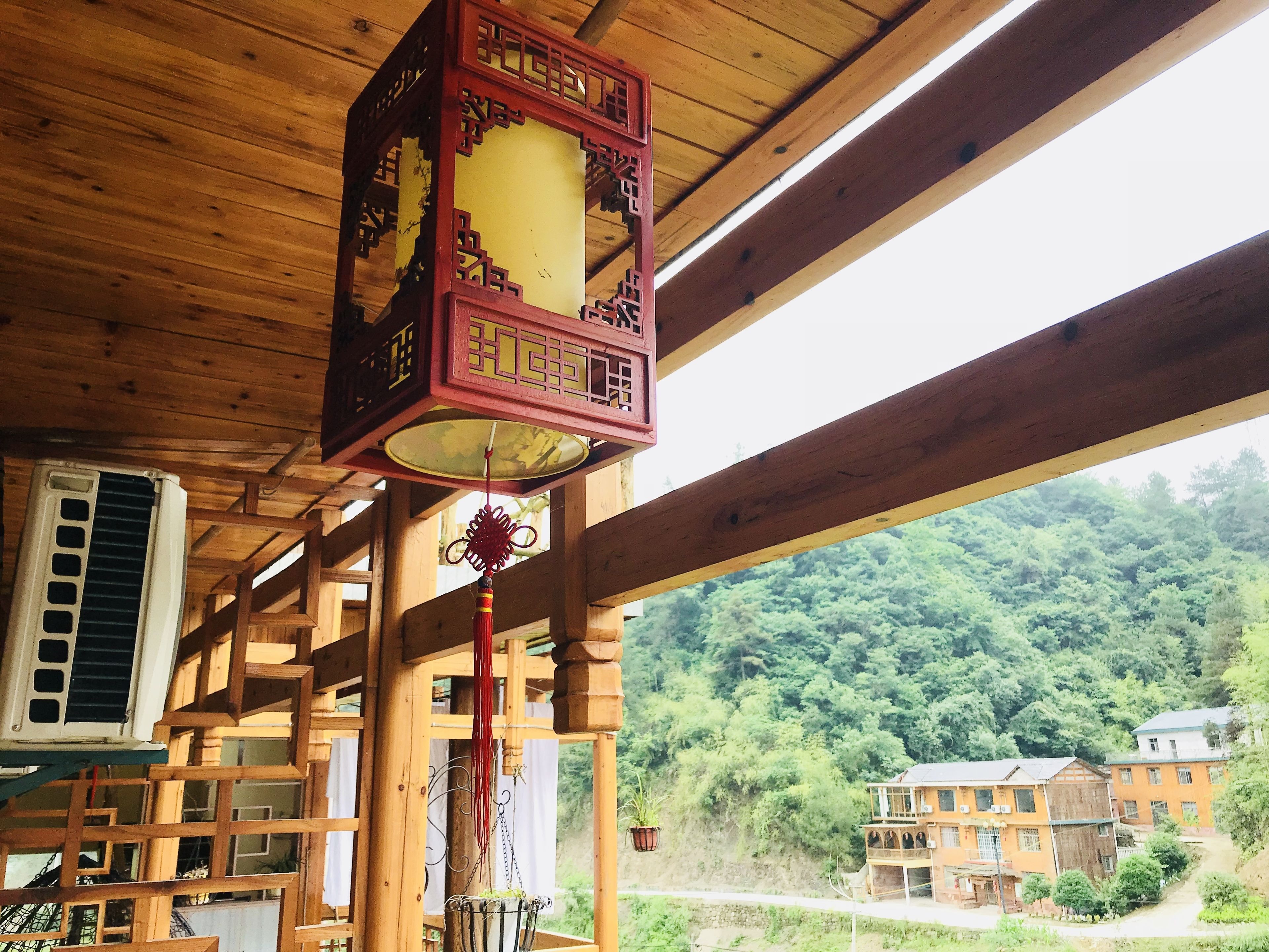 A wooden structure with a traditional Chinese lantern hanging from a beam, an air conditioning unit, and a view of a hillside with buildings.