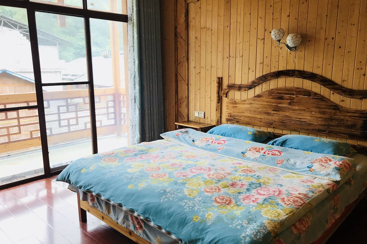 A wooden bed with floral bedding in a room with large windows and a view of a wooden deck outside.