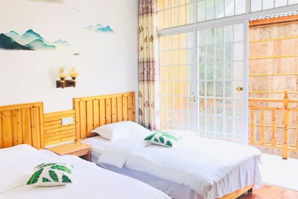 A room with two beds, wooden headboards, a wall with a mountain painting, and a large window with a view.