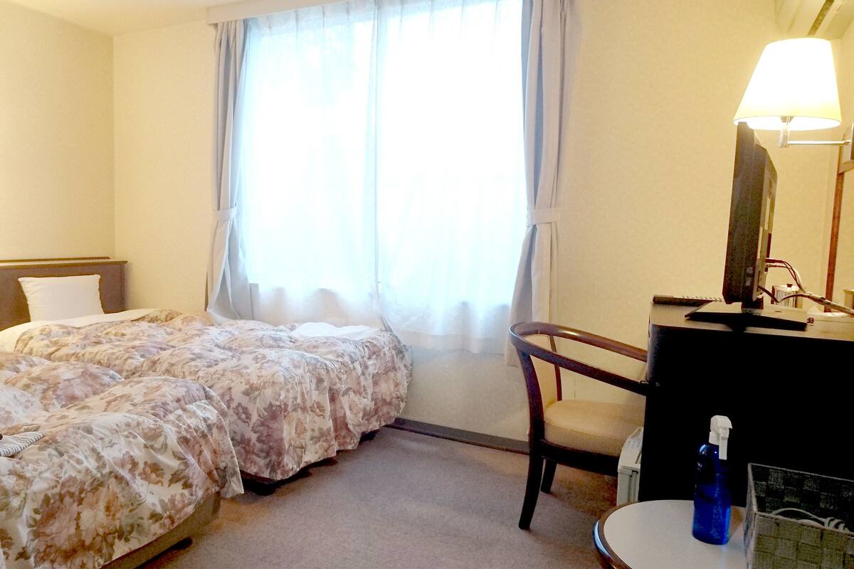 A hotel room with two beds, a desk, a chair, a lamp, and a window with curtains.