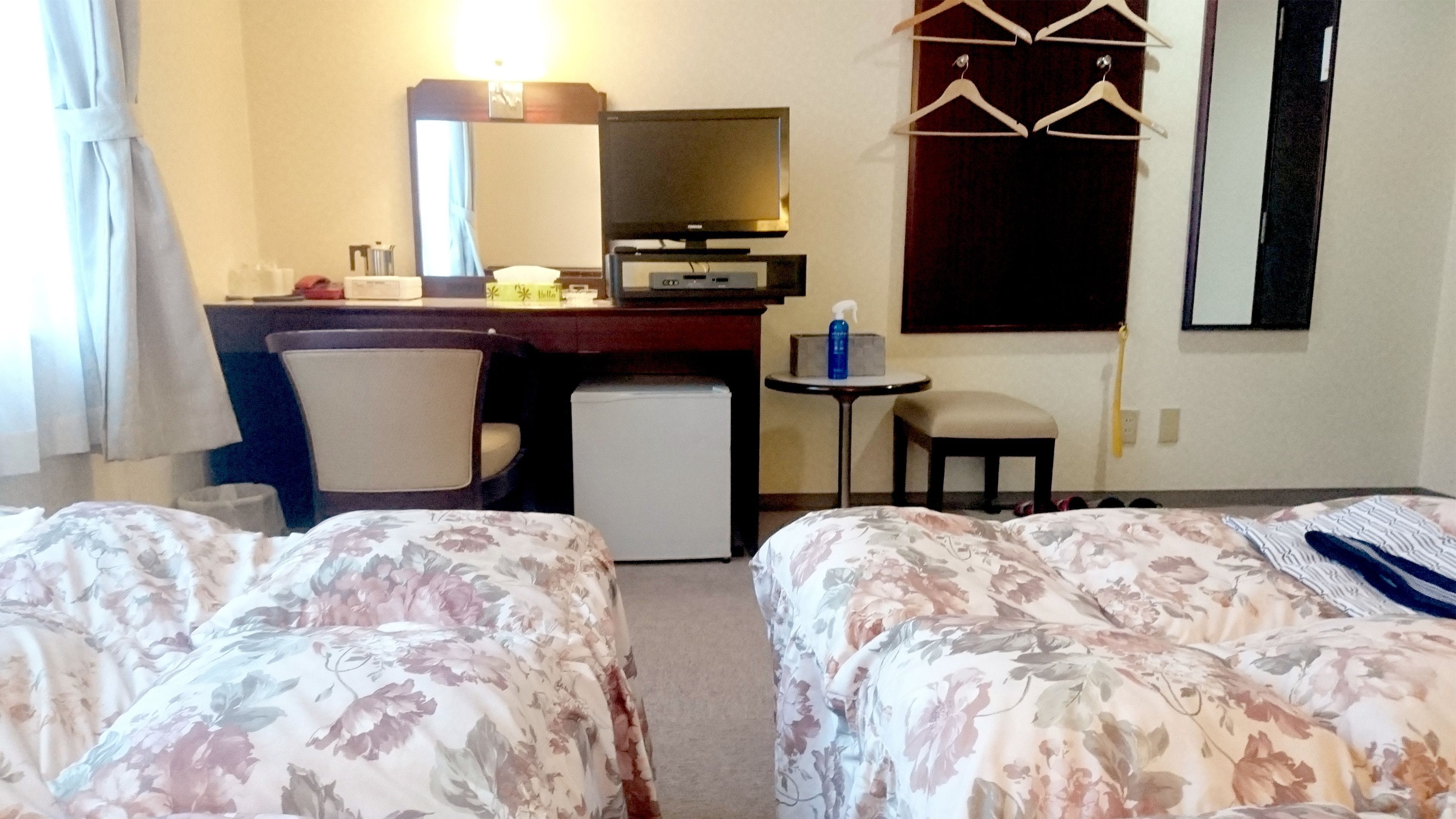 A hotel room with two beds, a desk with a computer, a small refrigerator, a chair, a table with a chair, and a TV.