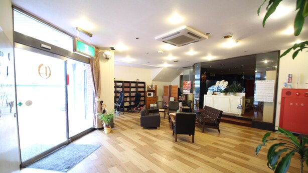 A spacious lobby with a wooden floor, a reception desk, a seating area with chairs, a bookshelf, and a potted plant. - Royal Inn Shinyamaguchi (Yamaguchi)