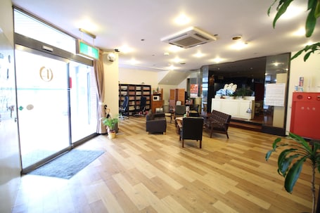 A spacious lobby with a wooden floor, a reception desk, a seating area with chairs, a bookshelf, and a potted plant.