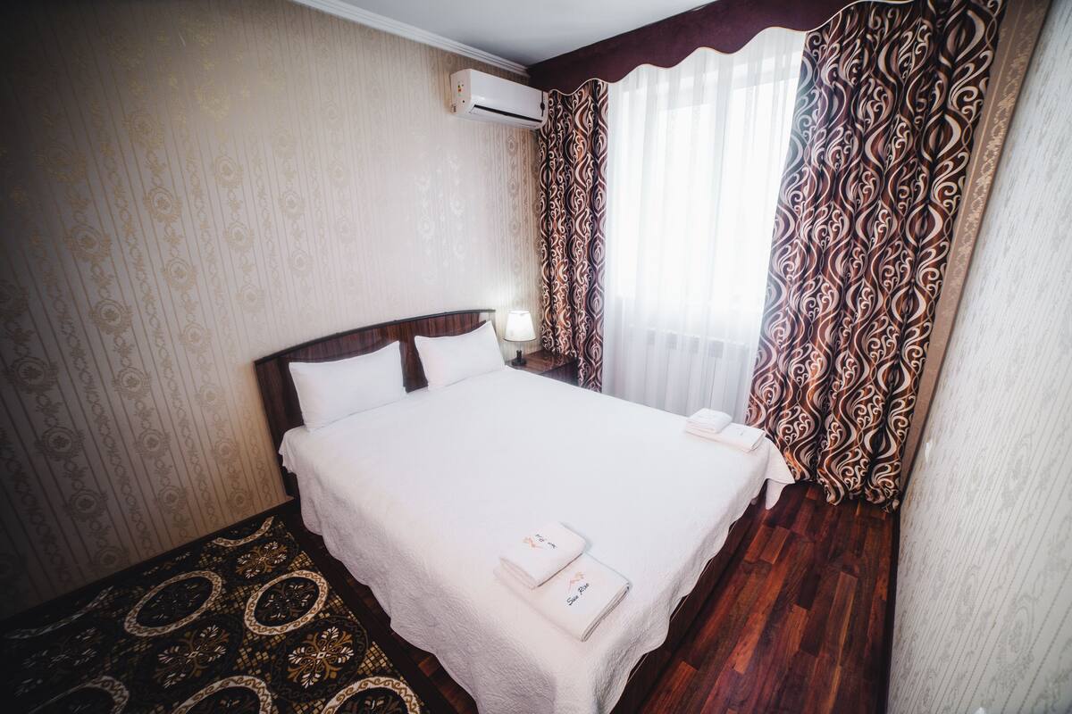 A hotel room with a neatly made bed, a nightstand with a lamp, patterned curtains, and a decorative rug.