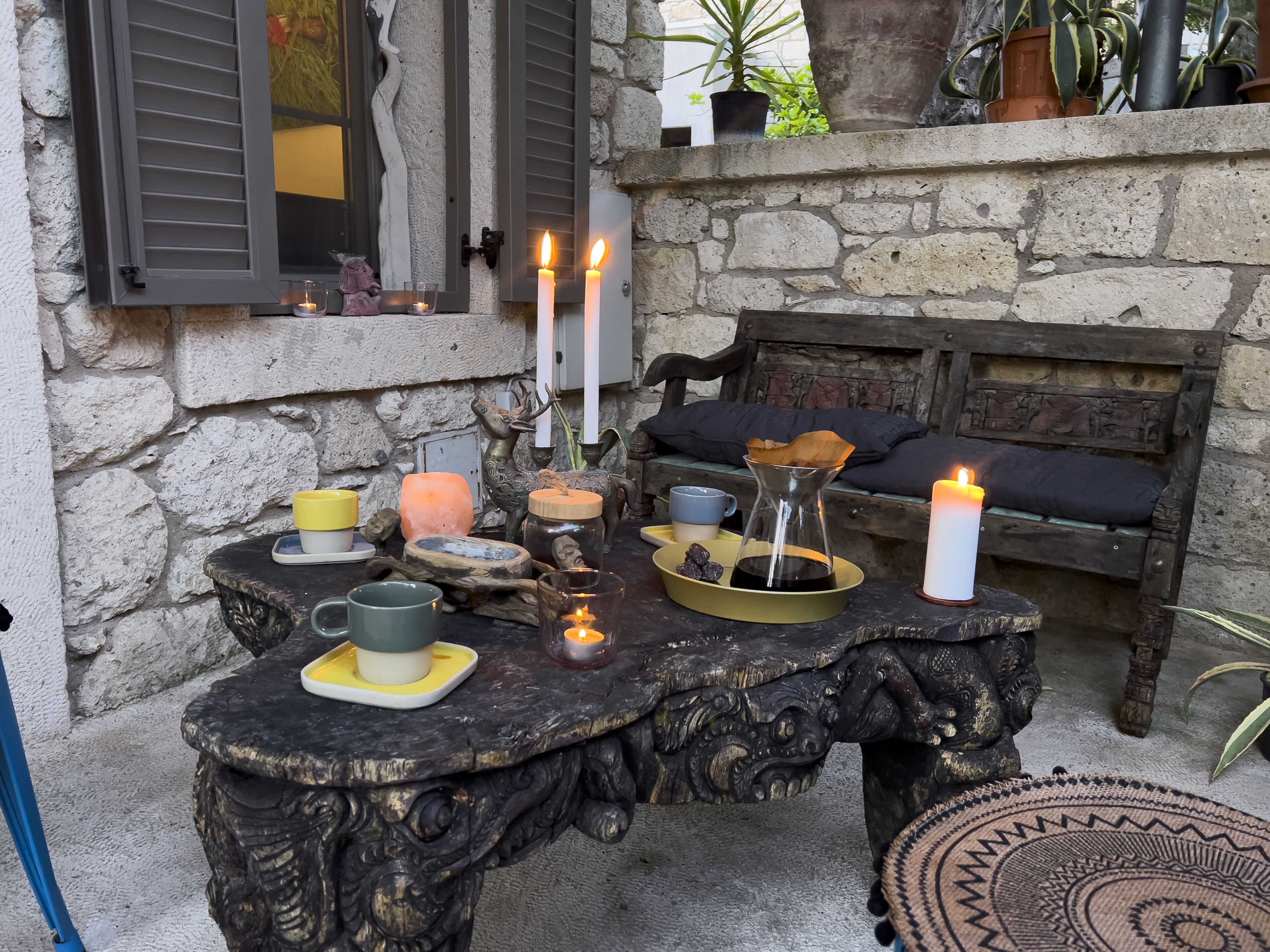 A rustic outdoor seating area with a stone wall, a wooden bench, and a table set with candles, a salt lamp, and various decorative items.