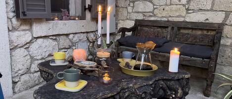 A rustic outdoor seating area with a stone wall, a wooden bench, and a table set with candles, a salt lamp, and various decorative items.