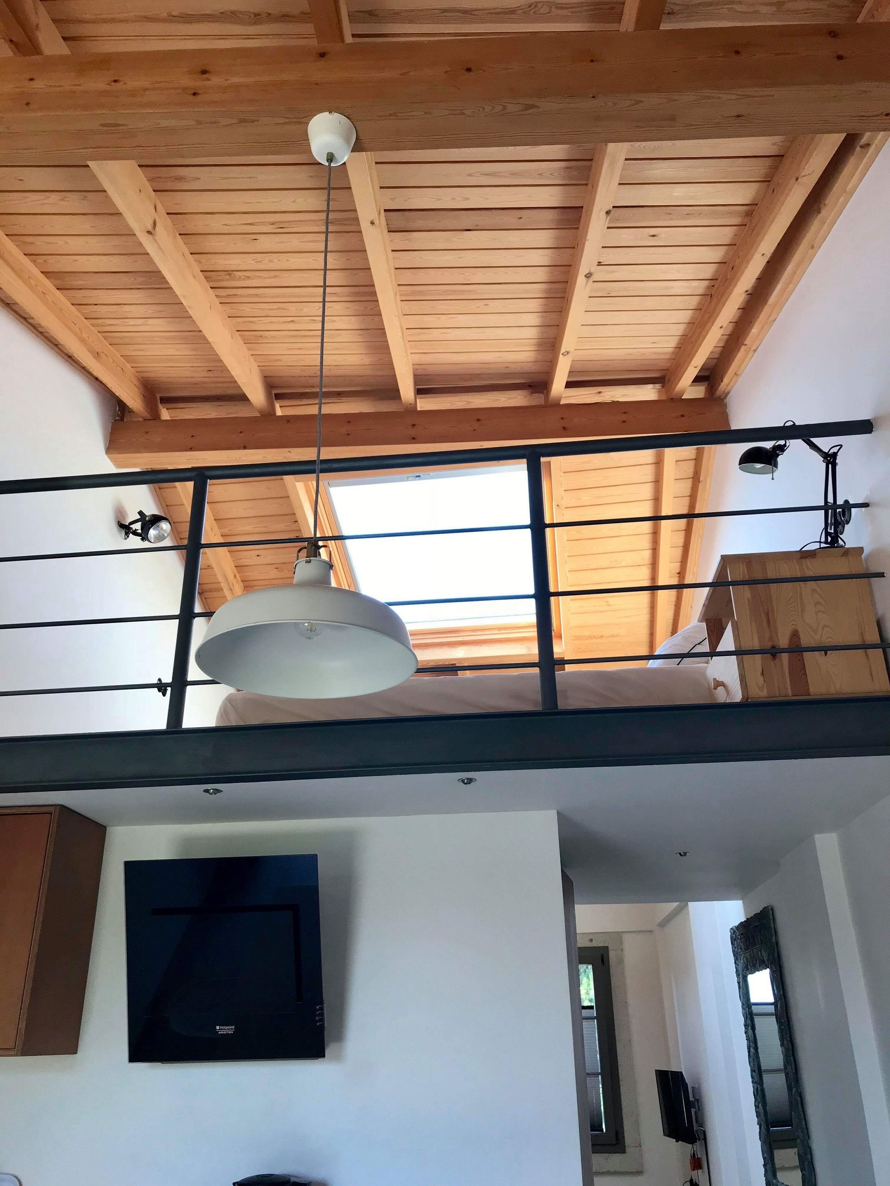 A room with a wooden ceiling, a balcony with a railing, a ceiling fan, and a mounted television.