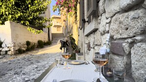 Mesa de comedor al aire libre dispuesta con platos, cubiertos y copas de vino, con vistas a una calle empedrada y un edificio de paredes de piedra.