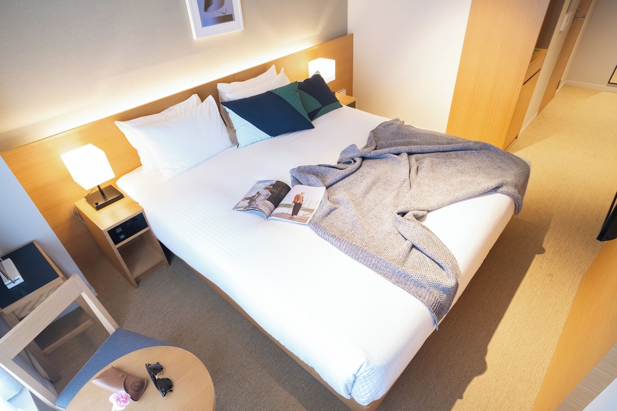 A neatly made bed with a blanket, pillows, and a magazine. There is a nightstand with a lamp and a small table with a handbag and a flower.