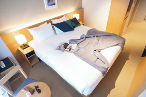 Una cama bien hecha con una manta, almohadas y una revista. Se ve un mesita de noche con una lámpara y una pequeña mesa con un bolso y una flor.