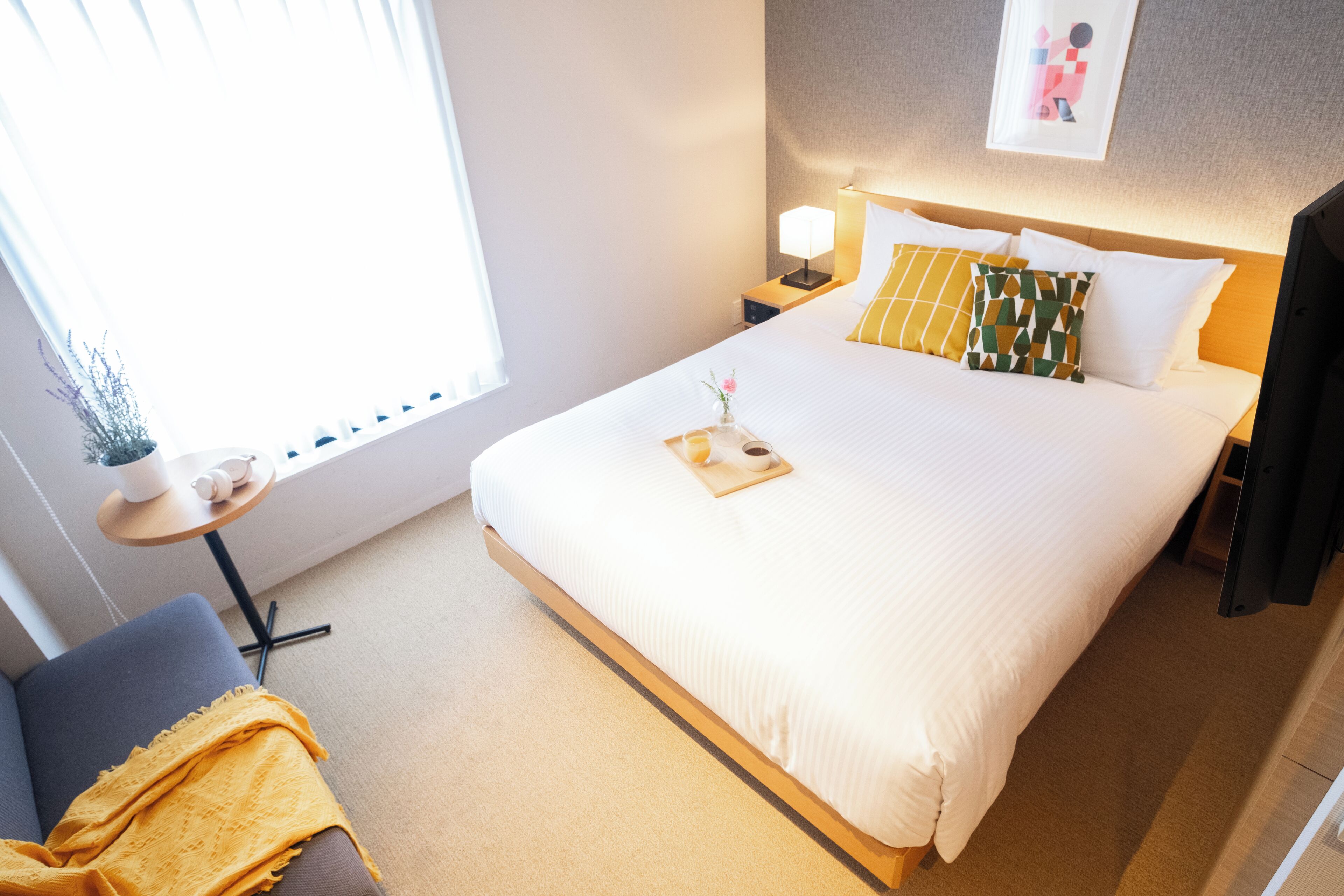 A hotel room with a large bed, a bedside table with a lamp, a small table with a plant, and a chair with a blanket.