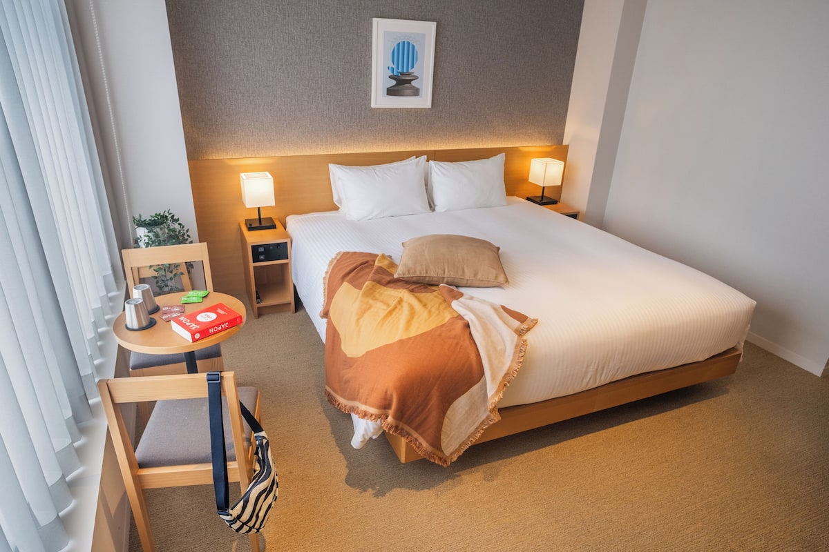 A hotel room with a bed, bedside lamps, a small table with a lamp, a chair, and a picture on the wall.