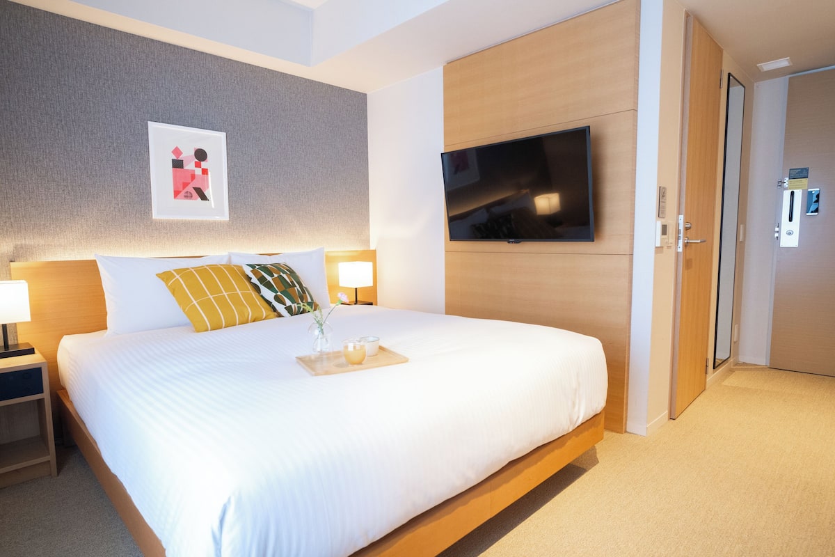A hotel room with a large bed, a flat-screen TV mounted on the wall, a bedside table with a lamp, and a door with a lock.