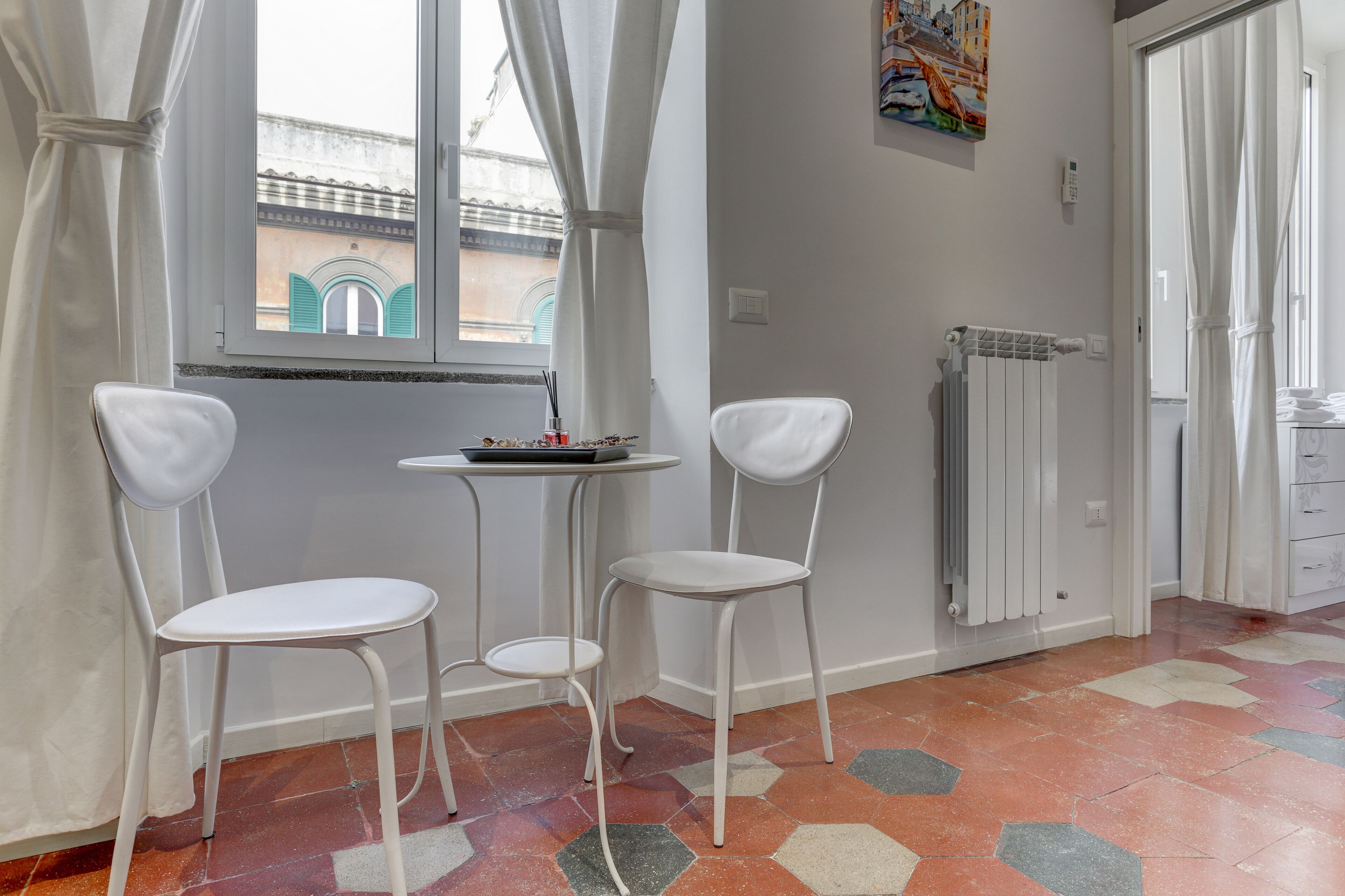 Une pièce avec deux chaises blanches, une petite table, un radiateur et une fenêtre donnant sur des immeubles.
