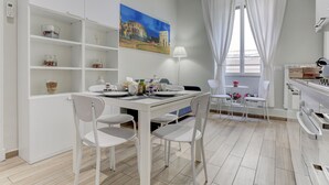 A modern dining area with a white table and chairs, a painting of a historical building, and a window with white curtains.