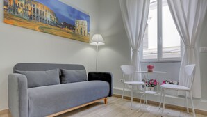 A modern room with a grey sofa, a white table with two chairs, a floor lamp, and a painting of a historical building.