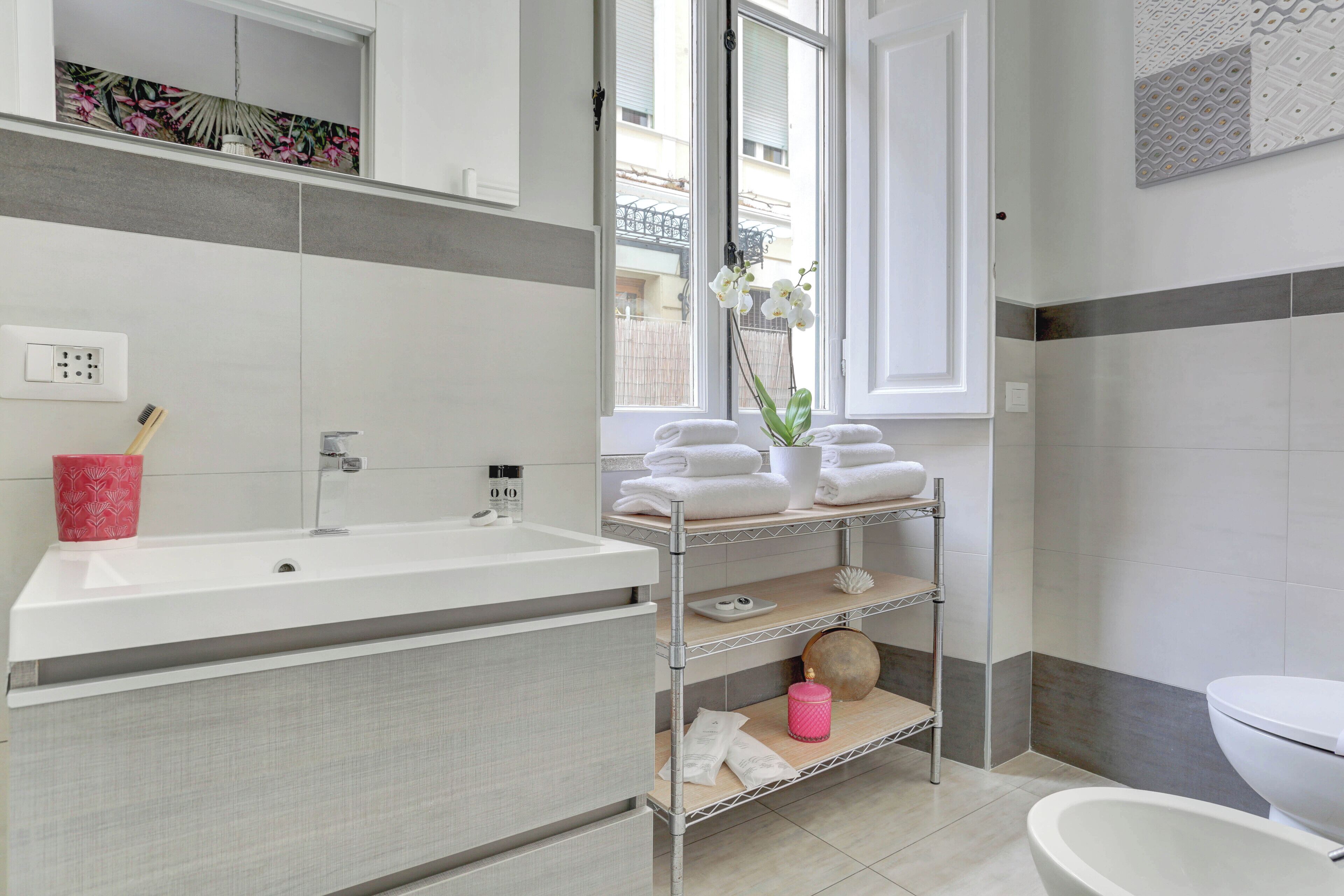 Ein modernes Badezimmer mit einem weißen Waschbecken, einer Toilette und einem Fenster mit Blick auf Gebäude.