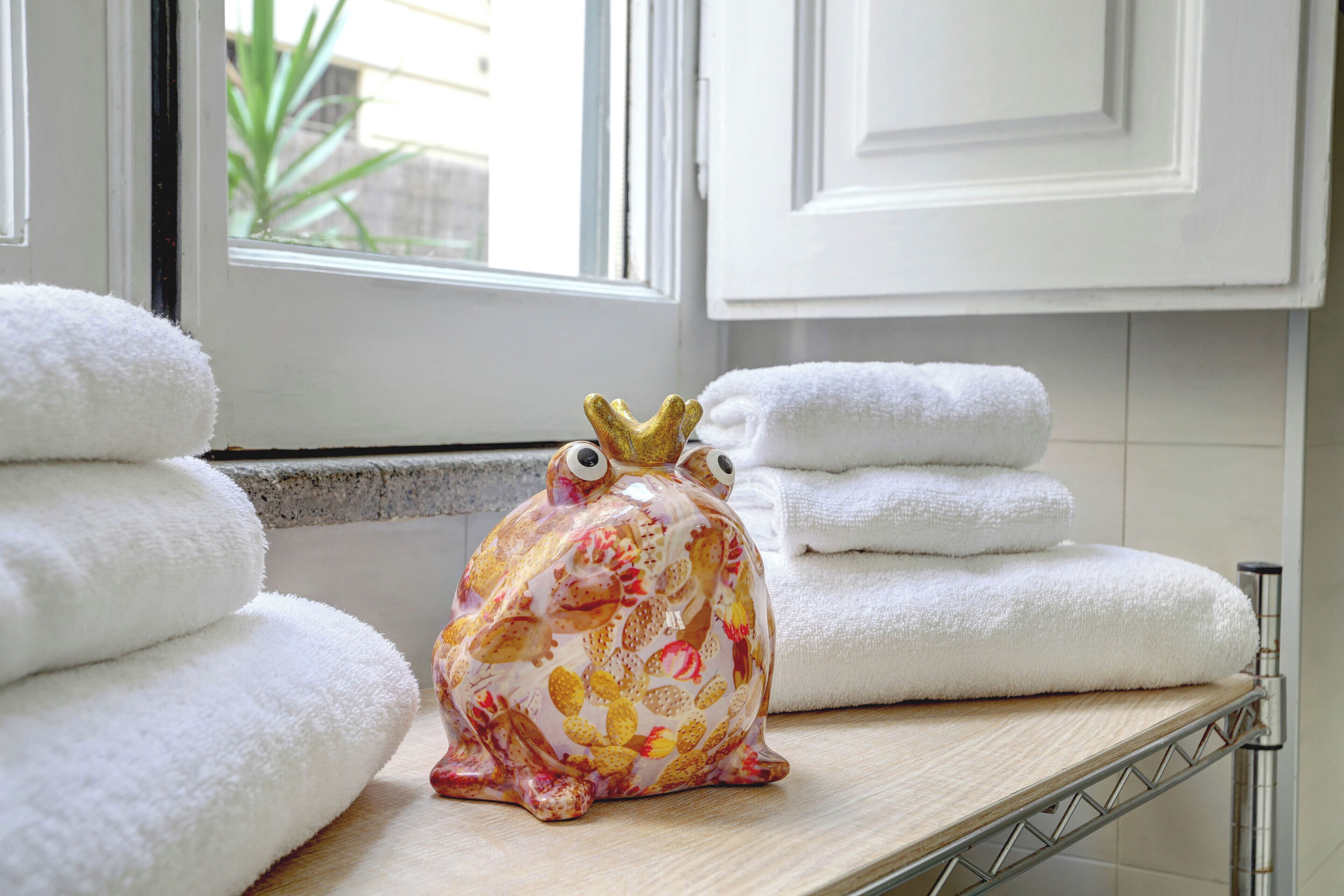 A bathroom with white towels stacked on a shelf, a decorative frog figurine, and a window with a view of greenery.