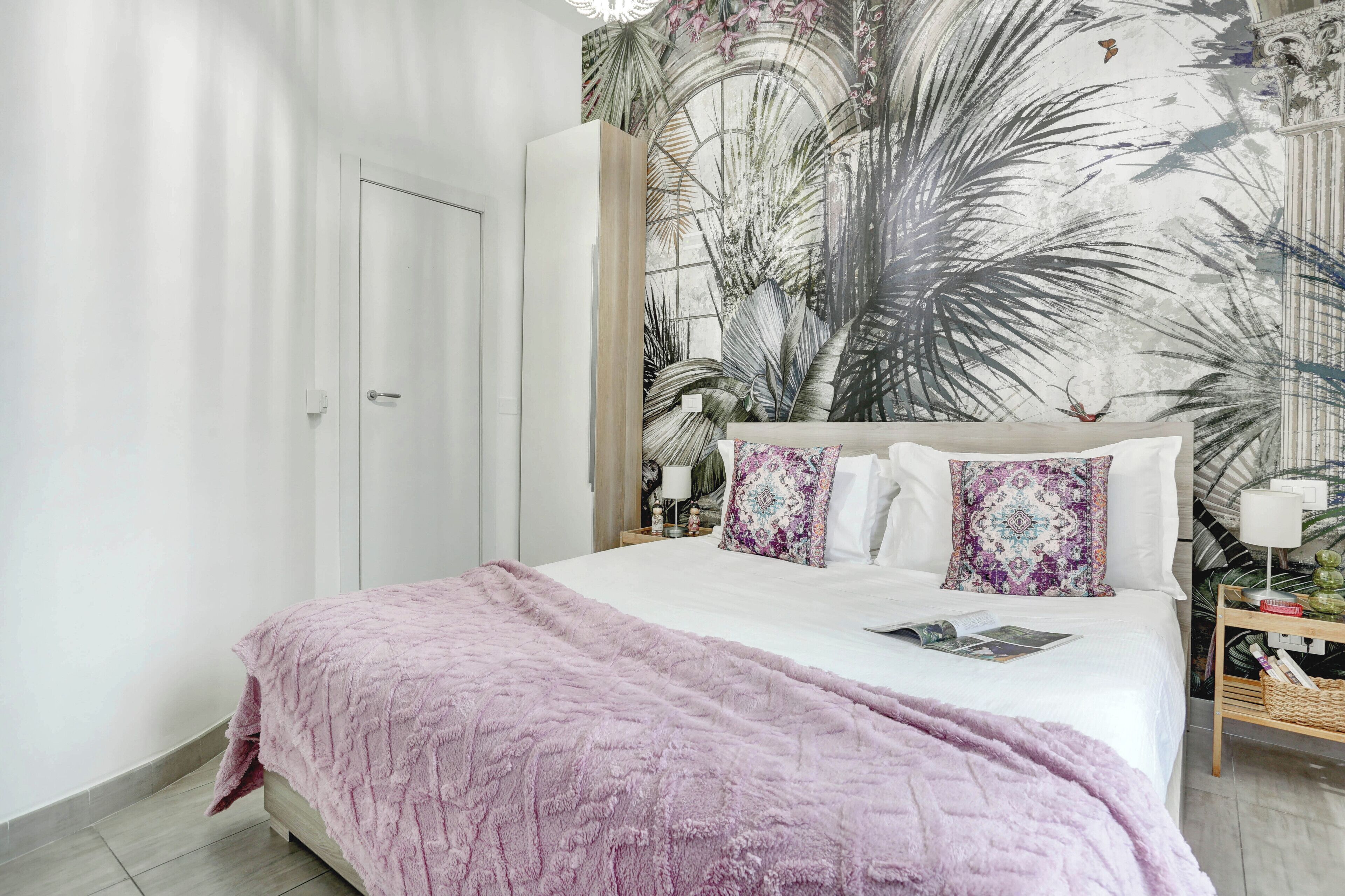 A bedroom with a bed, a nightstand, a door, and a wall with a tropical mural.