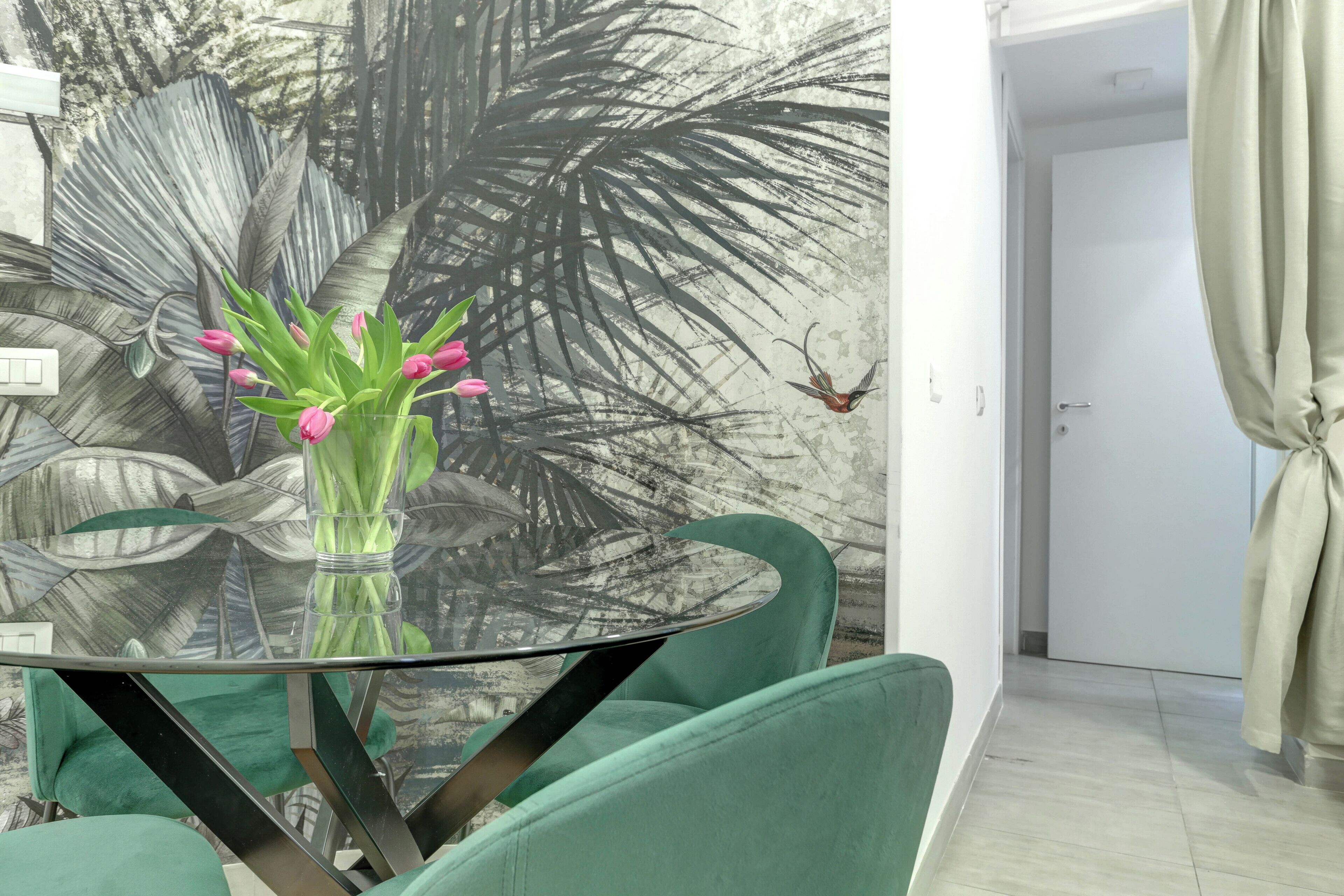 A dining area with a glass table, green chairs, and a wall mural of palm leaves.