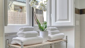 A bathroom with a towel rack holding folded towels, a potted plant, and a window with a view of a fence and buildings.