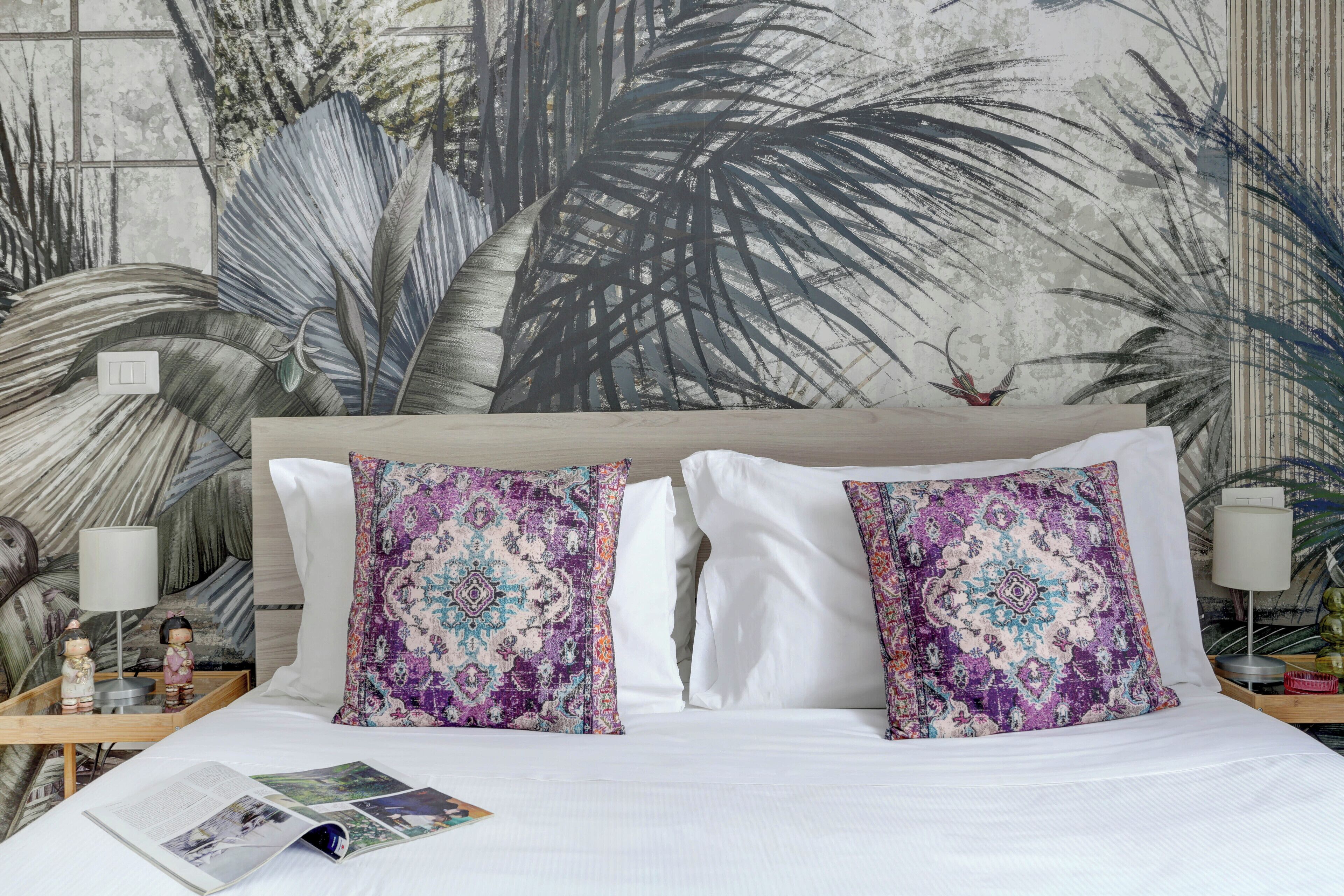 A neatly made bed with white linens, two patterned pillows, a bedside table with a lamp, and a wall mural of palm leaves.