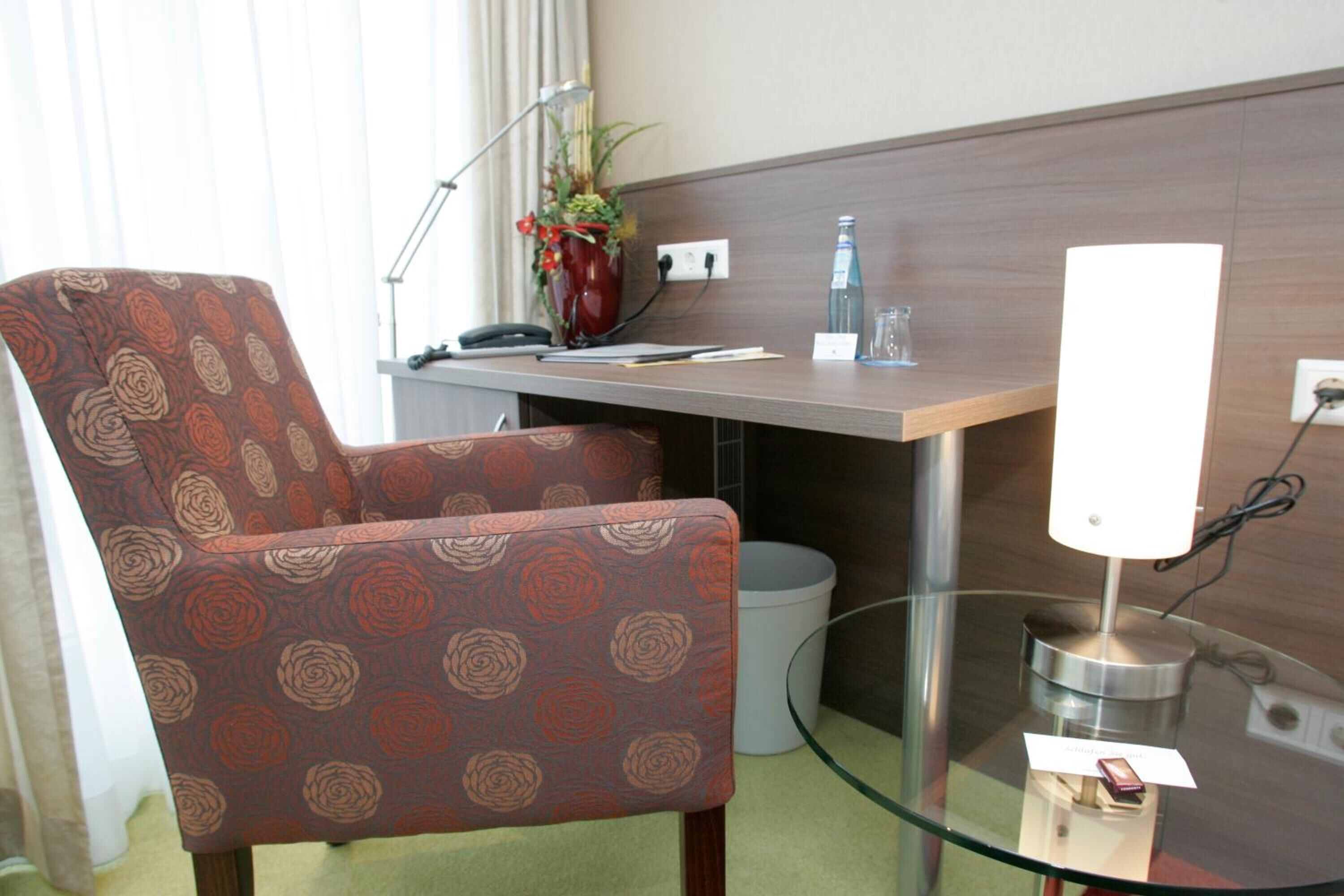 Une chambre d’hôtel avec un fauteuil à motifs, un bureau avec une lampe, un vase de fleurs et une table en verre avec une lampe.