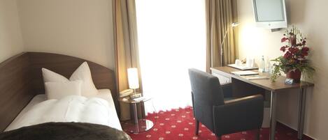 A hotel room with a bed, a desk, a chair, a TV, and a plant.