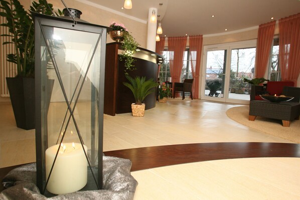 A reception area with a glass partition, a lit candle, potted plants, and a seating area with a sofa and chairs. - Isselhorster Landhaus (Guetersloh)