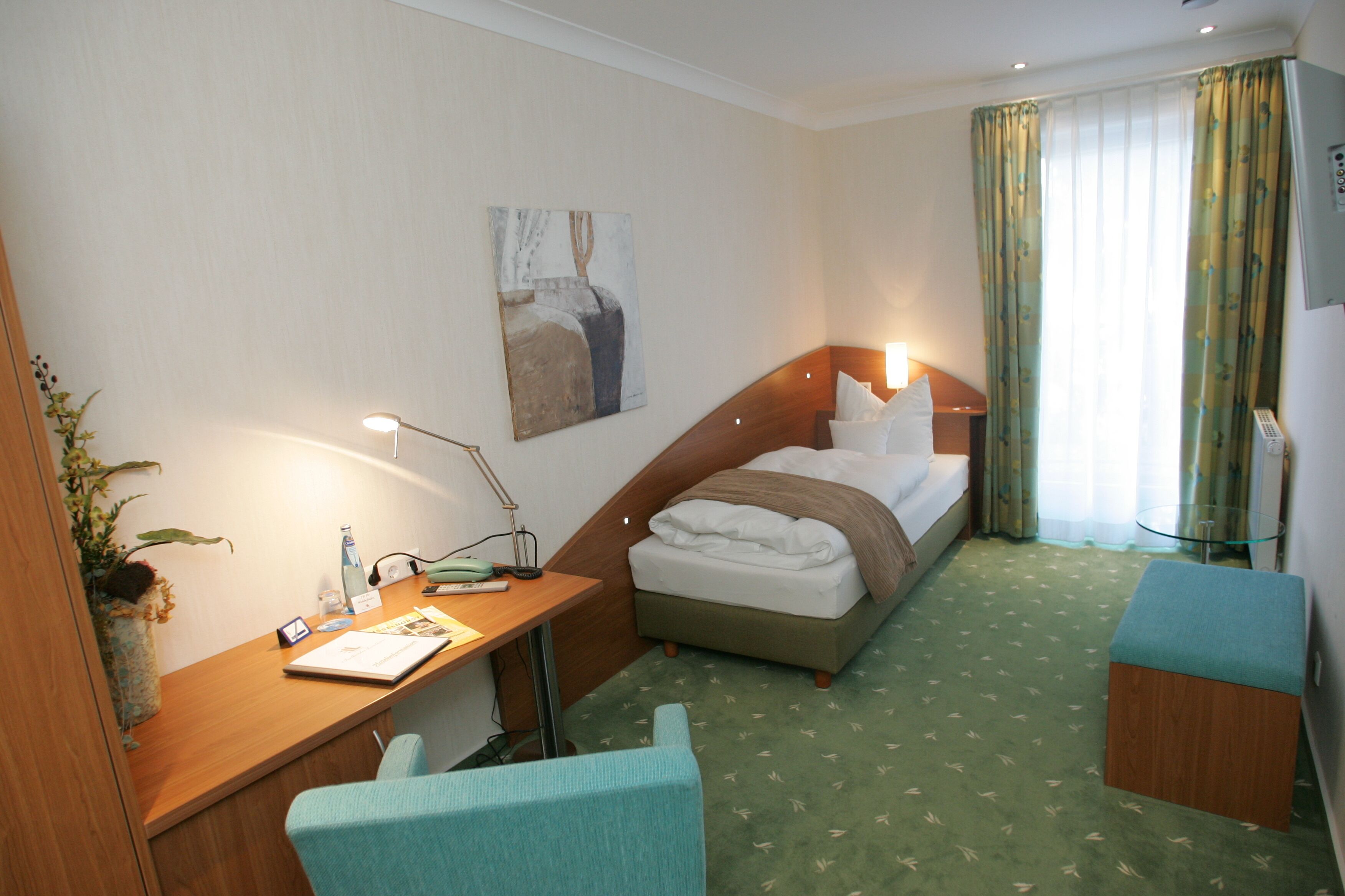 A hotel room with a bed, a desk with a lamp, a chair, a small table, and a painting on the wall.