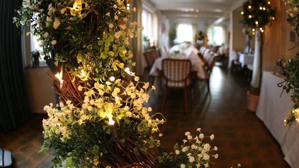 A restaurant interior with a long table and chairs, decorated with greenery and white flowers. - Isselhorster Landhaus (Guetersloh)
