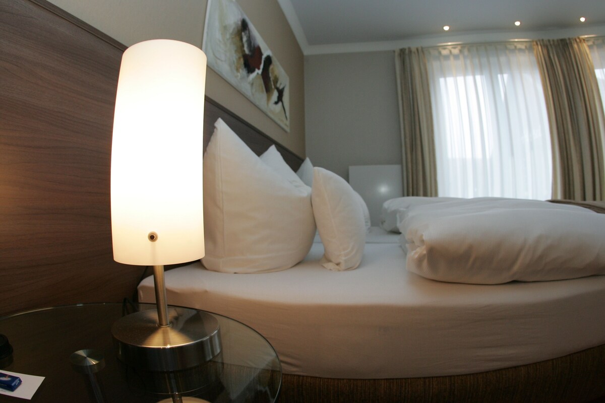 A hotel room with a bed, a bedside table with a lamp, and a painting on the wall.