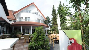A courtyard with outdoor seating, a white building with a red roof, and a painting of parrots. - Isselhorster Landhaus (Guetersloh)