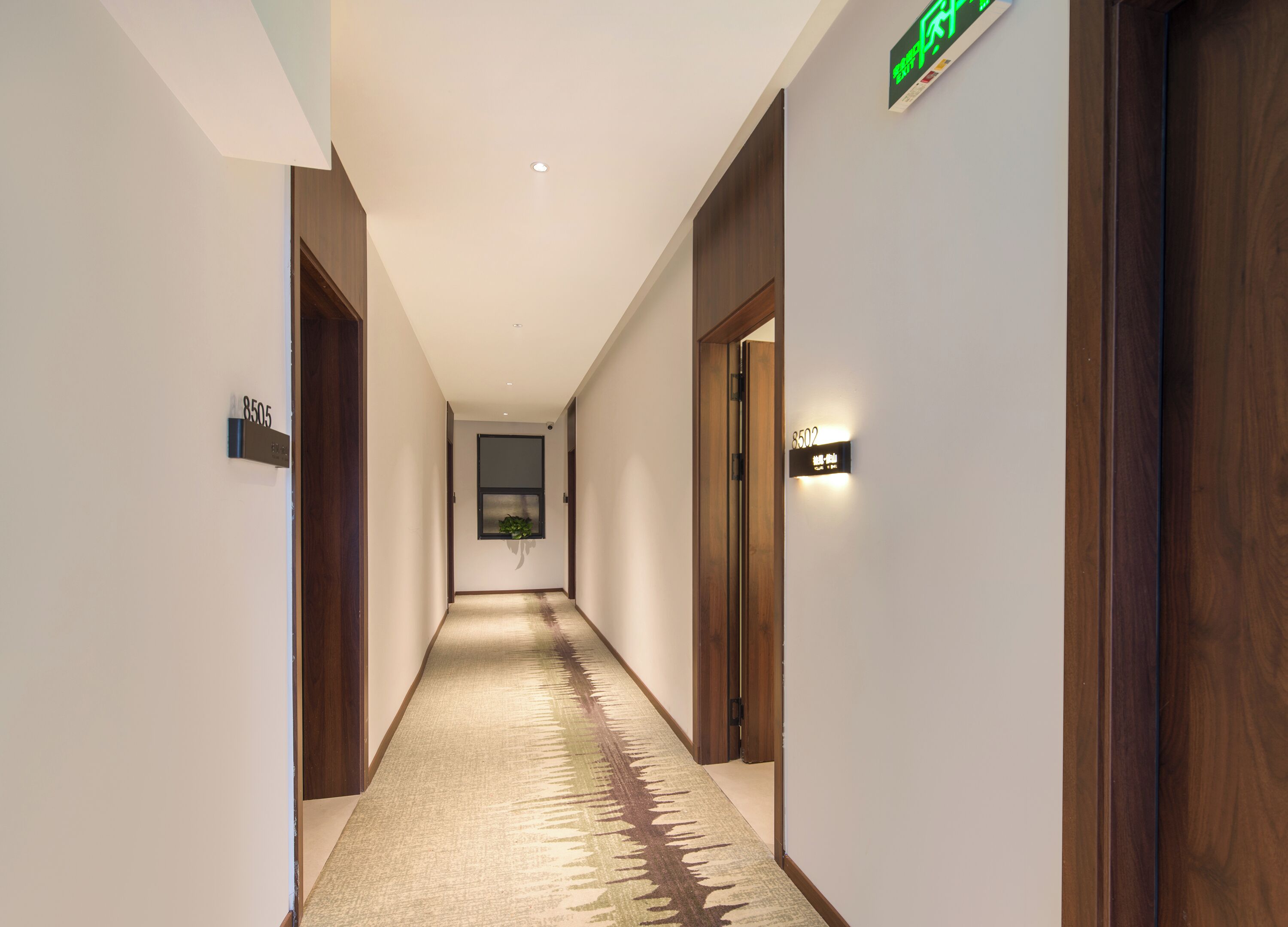 Un couloir d’hôtel avec des portes en bois, un sol recouvert de moquette et des panneaux lumineux indiquant les numéros de chambre.