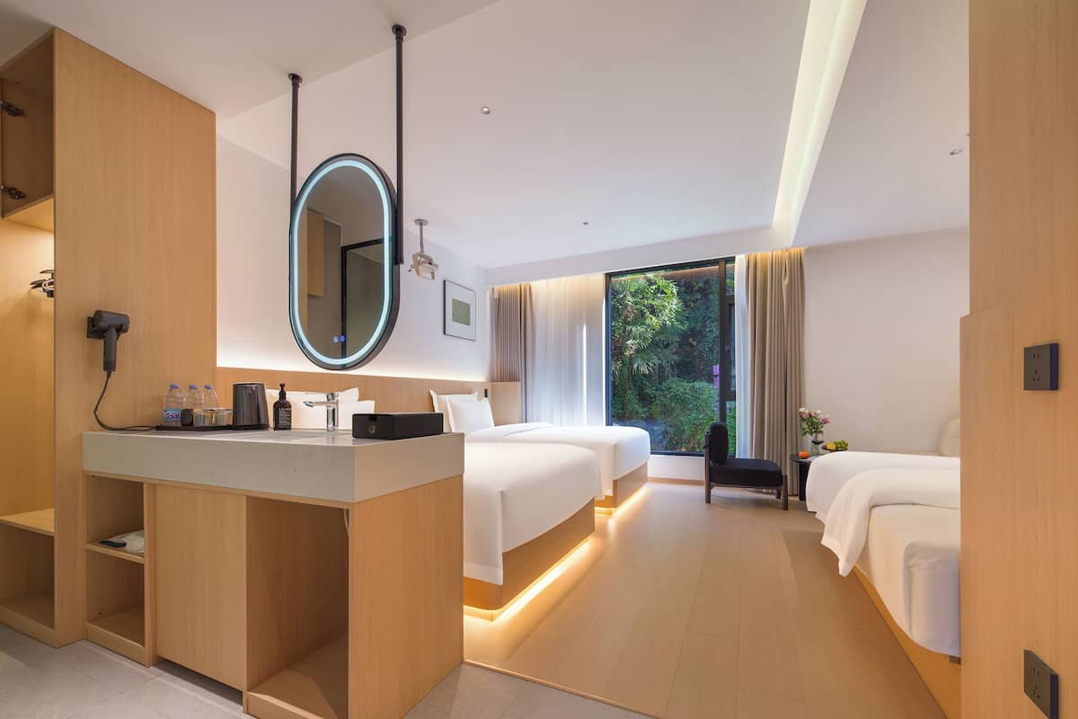 Habitación de hotel moderna con un escritorio de madera, un espejo grande, dos camas, una mesita y vistas a zonas verdes.