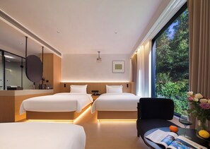 Ein Hotelzimmer mit zwei Betten, einem großen Fenster mit Blick auf Grünflächen, einem Nachttisch mit Buch und Glas sowie einem Spiegel.