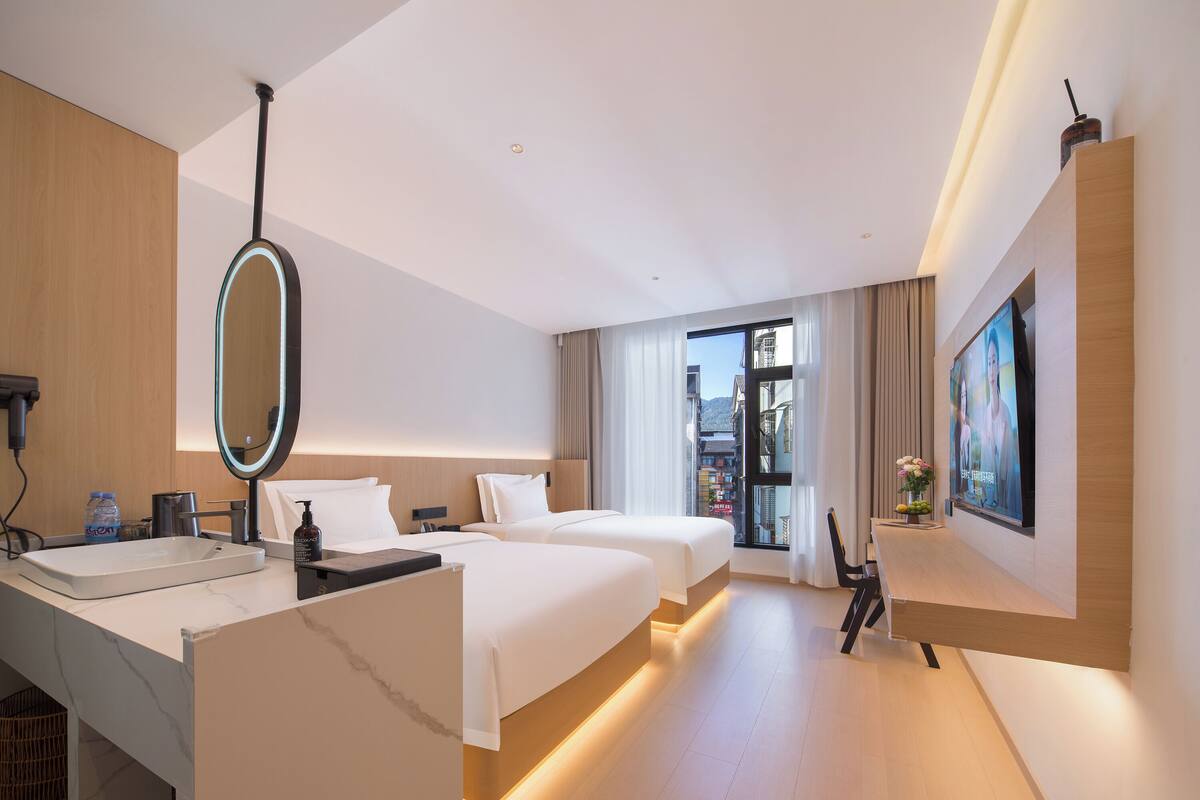 Una habitación de hotel moderna con una cama grande, una cama más pequeña, un escritorio con espejo, un televisor y una ventana con vistas a la ciudad.