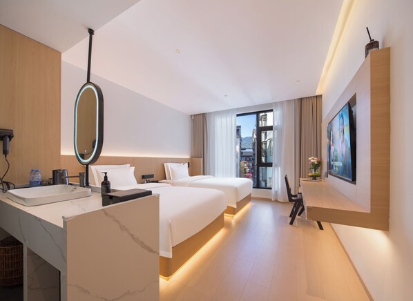 Ein modernes Hotelzimmer mit einem großen Bett, einem Einzelbett, einem Schreibtisch mit Spiegel, einem Fernseher und einem Fenster mit Blick auf die Stadt.