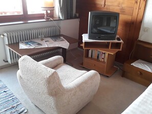 Una habitación pequeña con un radiador, un escritorio de madera con lámpara, un televisor sobre un mueble, un sillón blanco y una estantería.