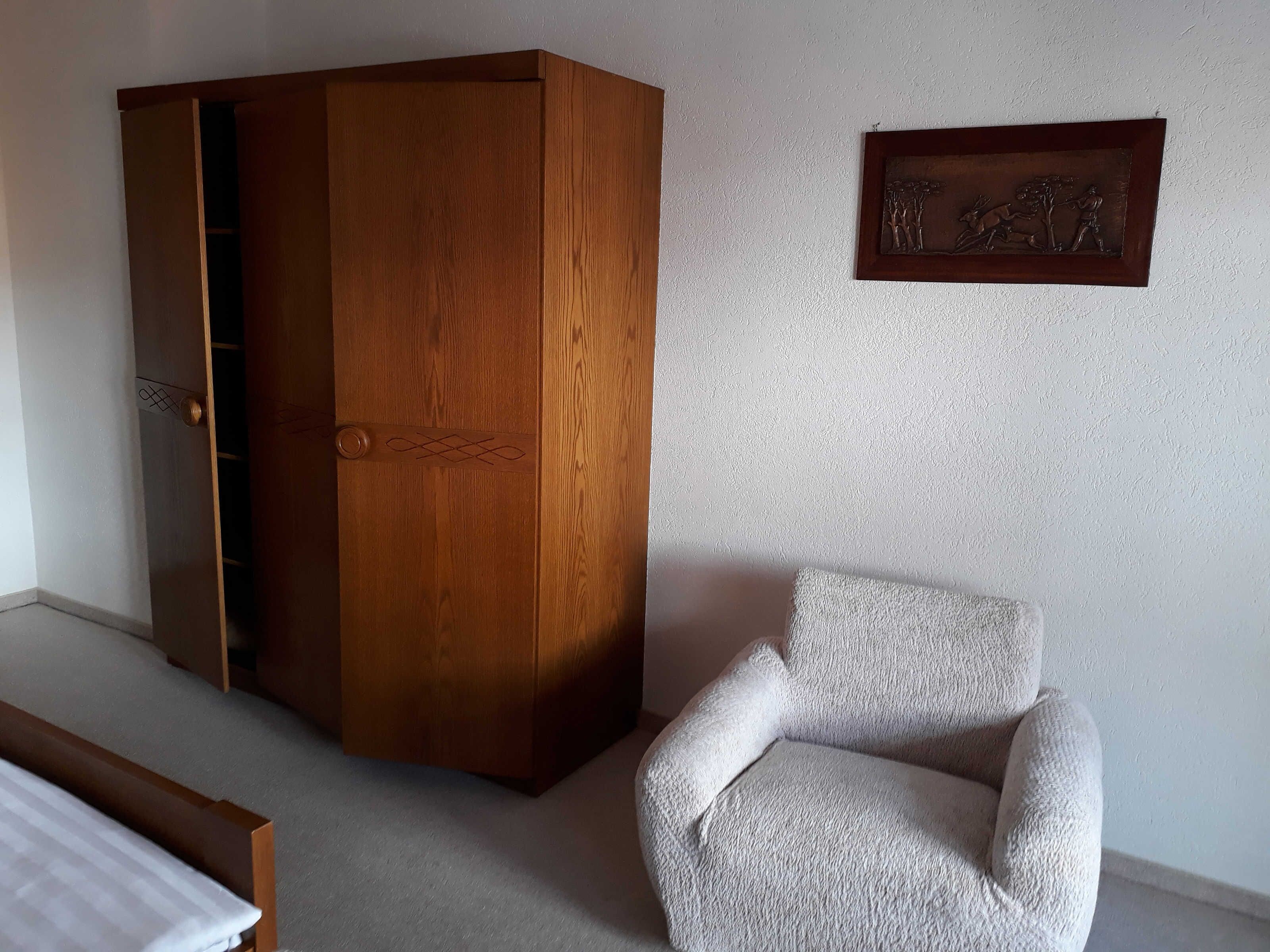 Een kamer met een houten garderobe, een witte fauteuil en een kunstwerk in een lijst aan de muur.