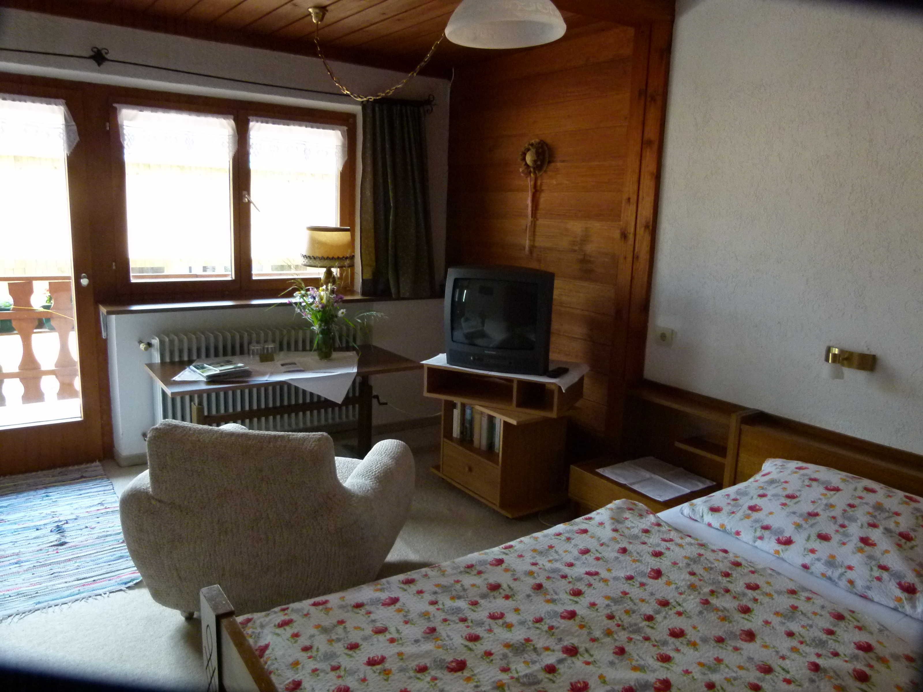 Een kamer met een bed, een stoel, een klein tafeltje met een lamp, een televisie op een standaard en een raam met gordijnen.