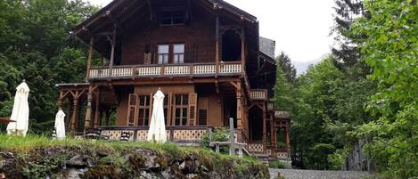 A large wooden house with a balcony, surrounded by greenery.