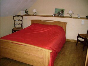 A bedroom with a wooden bed, a red bedspread, a wooden chair, a nightstand with a lamp, and a shelf with decorative items. - La Guguenaie (Beganne)