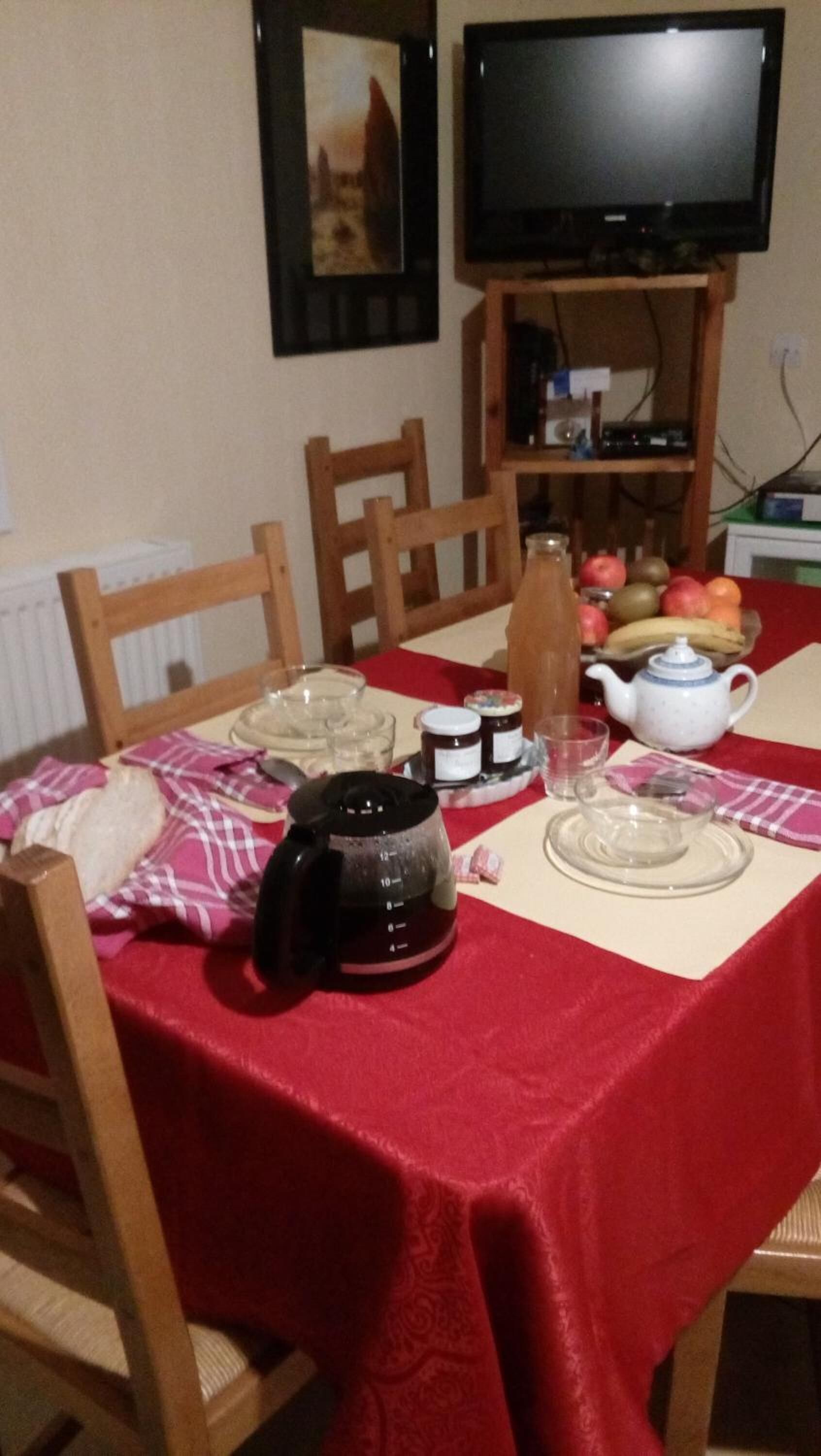 A dining table with a red tablecloth, wooden chairs, a teapot, jars, a glass pitcher, and a bowl of fruit.