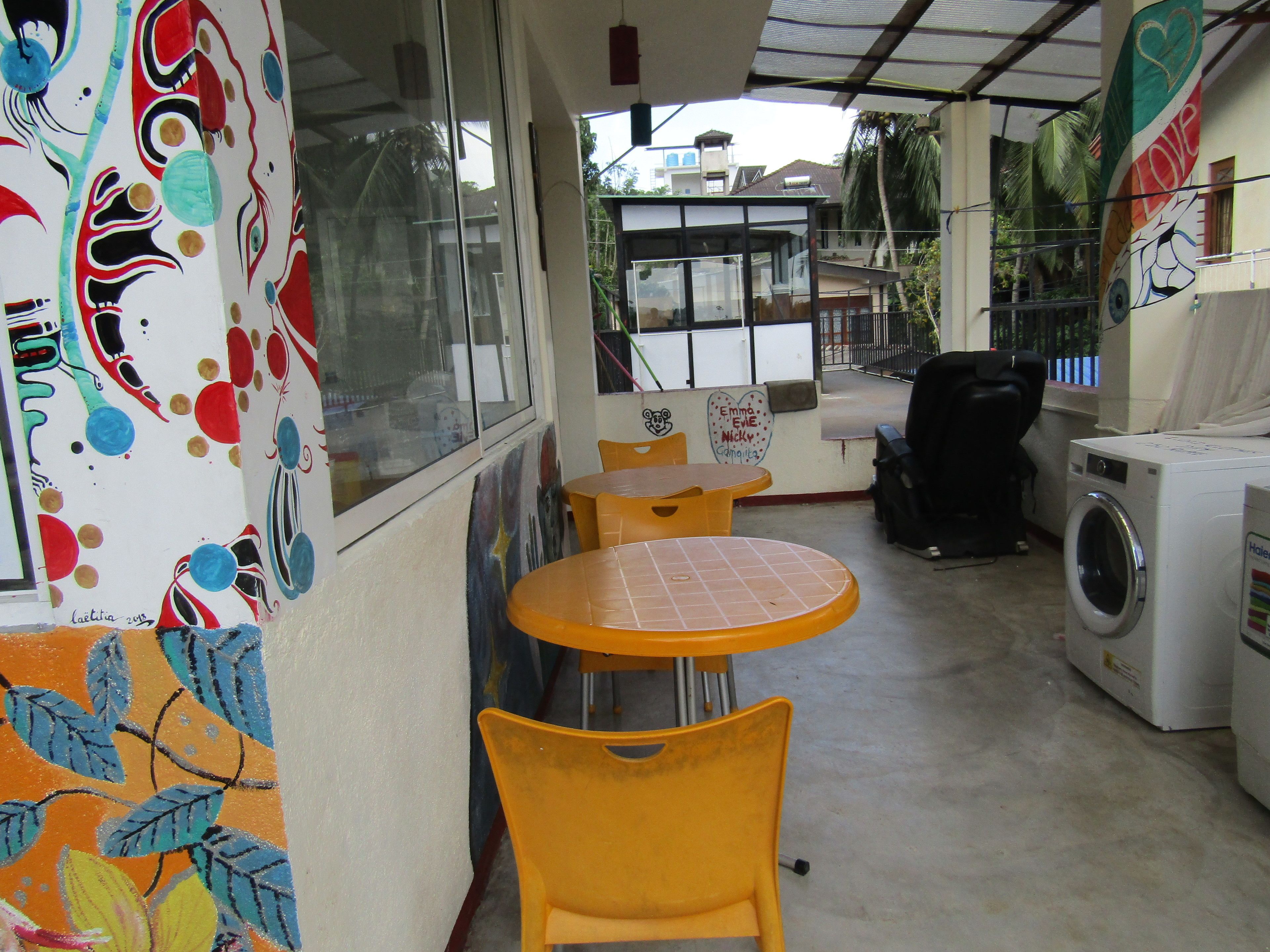 A small, enclosed laundry area with a washing machine, two yellow plastic chairs, and a window with a view of buildings outside.