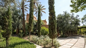 A garden with a paved walkway, tall cypress trees, and a building with a red roof. - Villa Touareg Domaine des Kasbahs (Marrakech)