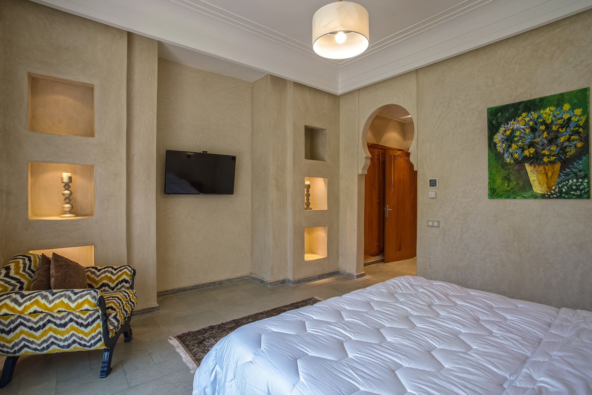 A bedroom with a large bed, a TV mounted on the wall, and a painting of flowers.